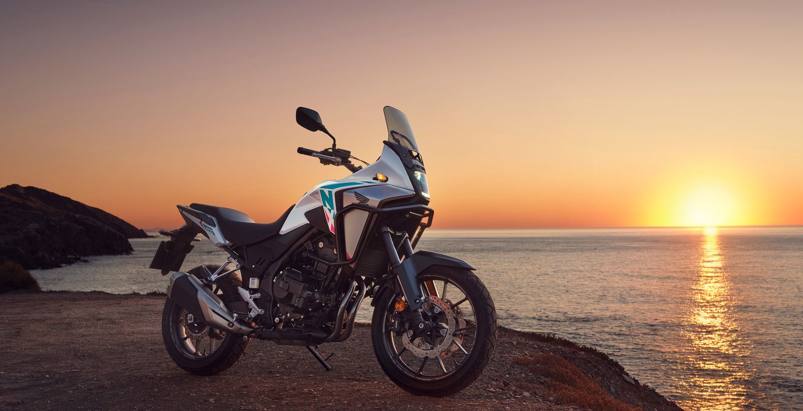Une Honda NX500 blanche garée au bord d'une falaise, face à la mer au coucher du soleil.